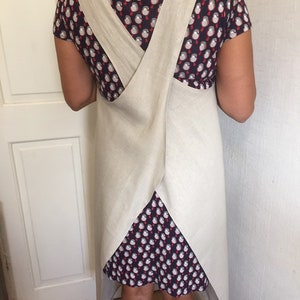 May include: A beige linen apron with criss-cross straps at the back. The apron is long and covers the wearer's entire back.