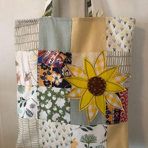 May include: A patchwork tote bag featuring a sunflower applique. The bag is made from various fabrics with floral and geometric patterns in shades of green, yellow, and brown. The sunflower is yellow with a brown centre and has a checkered pattern.