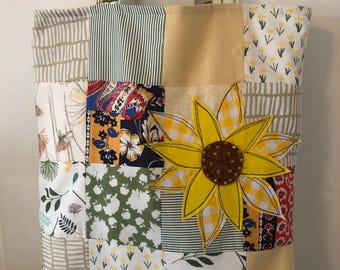 May include: A patchwork tote bag with a sunflower applique. The bag is made of different fabrics with floral and geometric patterns in shades of green, yellow, and brown. The sunflower is yellow with a brown center and has a checkered pattern.