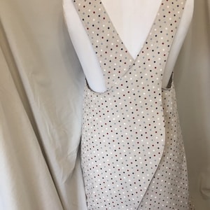 May include: A beige apron with a criss-cross back strap. The apron has a white background with small red, blue, and grey polka dots.