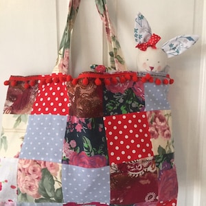 May include: A patchwork tote bag with red polka dot fabric, floral fabric, and blue fabric with white hearts. The bag has red pom pom trim and a white bunny with red bow is peeking out of the top.