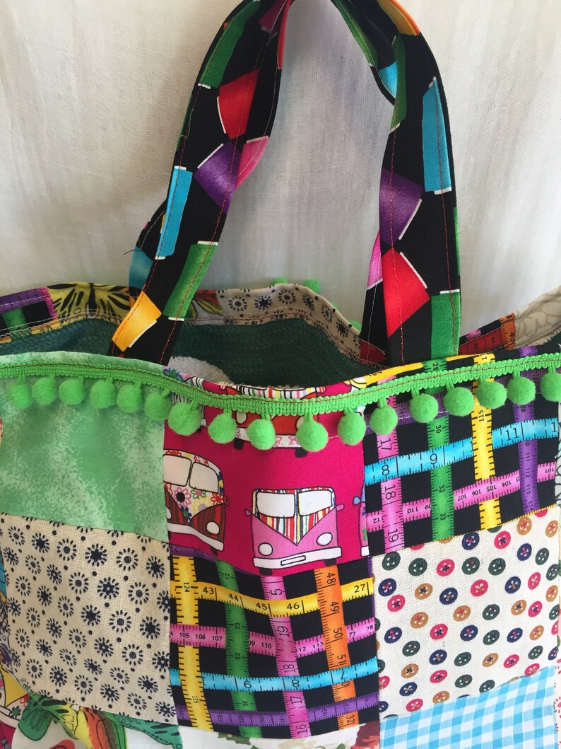 May include: A patchwork tote bag with green pom pom trim. The bag is made of different fabrics, including a green floral print, a pink fabric with a pattern of colorful vans, a black and white fabric with a ruler pattern, and a beige fabric with a pattern of colorful buttons. The bag has two black handles with a colorful geometric pattern.