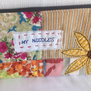 May include: A patchwork fabric zippered pouch with a yellow flower applique and a white label that says "MY NEEDLES".