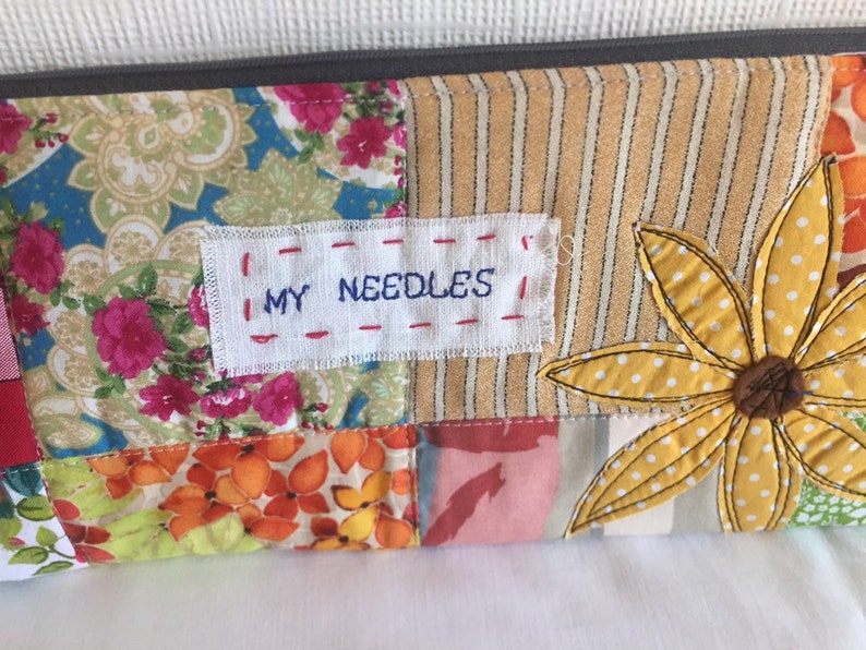 May include: A patchwork fabric zippered pouch with a yellow flower applique and a white label that says "MY NEEDLES".
