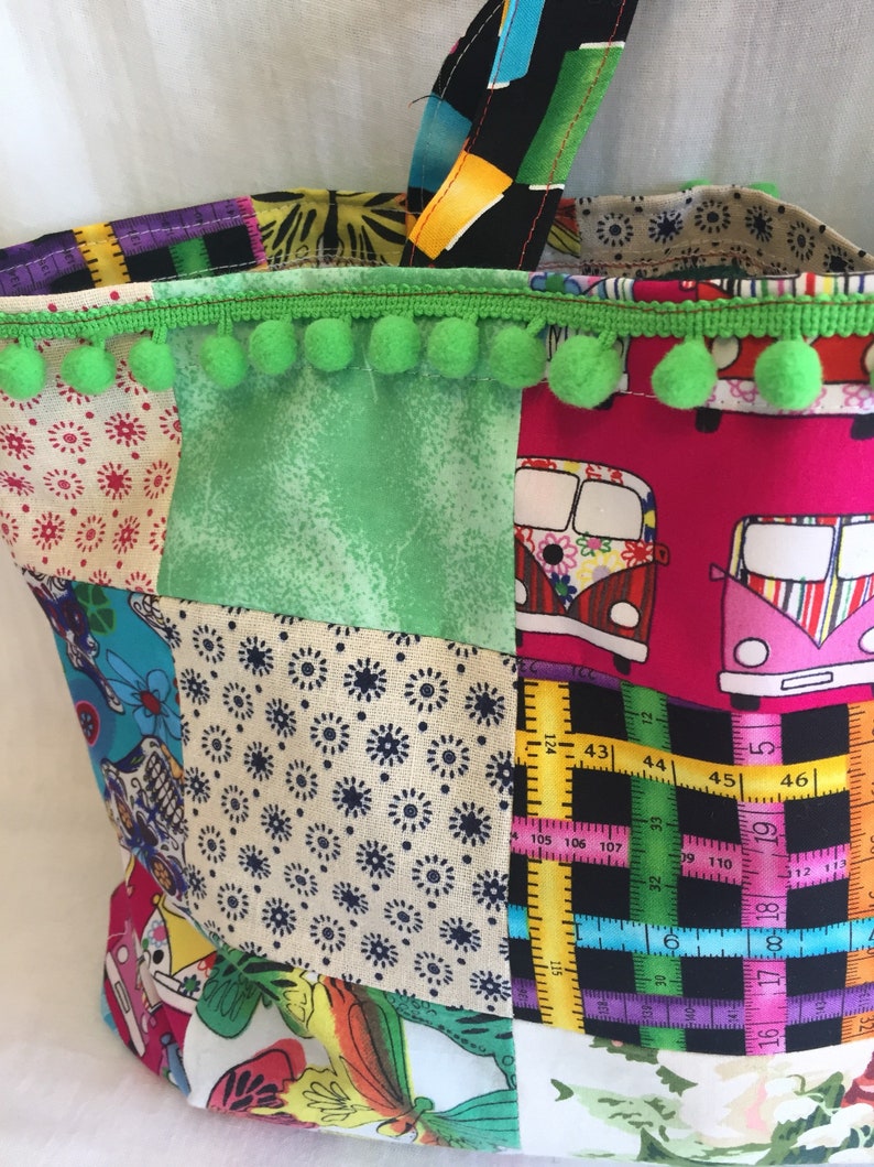 May include: A patchwork tote bag with a green pom-pom trim. The bag is made of different fabrics with various patterns, including flowers, polka dots, and a ruler design. The bag has a black and white fabric handle.