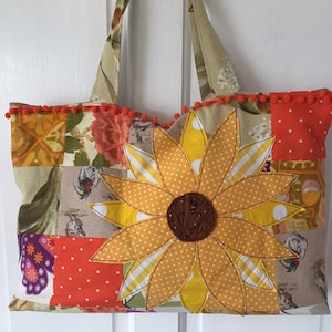 May include: A patchwork tote bag with a large yellow sunflower appliqué. The bag has a brown fabric handle and a white pom-pom trim.