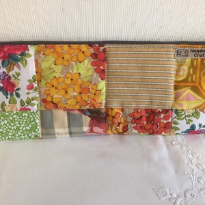 May include: A patchwork fabric zippered pouch with a variety of floral and geometric patterns in shades of yellow, orange, red, green, and brown. The pouch has a brown zipper and a label that reads "Handmade By CraftyHe."