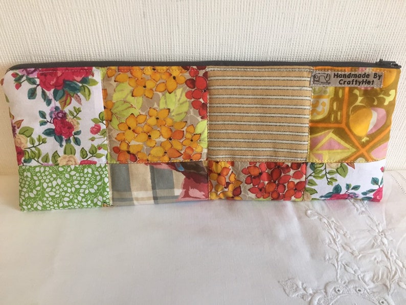 May include: A patchwork fabric zippered pouch with a variety of floral and geometric patterns in shades of yellow, orange, red, green, and brown. The pouch has a brown zipper and a label that reads "Handmade By CraftyHe."