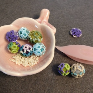 May include: A collection of colorful crocheted beads in a pink bowl. The beads are in shades of blue, green, purple, and white. There are also some white beads scattered around the bowl.