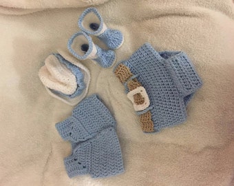 Puede incluir: Un conjunto de bebé hecho a mano en crochet azul claro y blanco. El conjunto incluye botines, un gorro, un cubrepañales con hebilla beige y calentadores de piernas. Los artículos están dispuestos sobre una superficie blanca y suave.