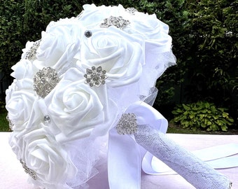 May include: A white satin bridal bouquet with a lace-covered handle and a white satin ribbon. The bouquet is decorated with silver rhinestone brooches.