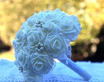 May include: A white bridal bouquet made of artificial roses with pearl accents. The bouquet is wrapped in white tulle and has a white satin ribbon.