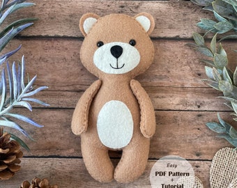 May include: A brown felt teddy bear with a white felt belly and black eyes. The bear is standing on a wooden surface with a sign that says "Easy PDF Pattern + Tutorial".