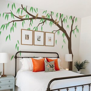 May include: A white bed with a black metal frame and white bedding. The bed has a white headboard and footboard. There are three framed pictures on the wall behind the bed. A green and brown tree decal is on the wall above the bed. There is a small table next to the bed with a lamp on it.