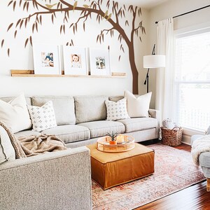 May include: A cozy living room with a light gray sofa, a brown leather ottoman, and a patterned rug. Three framed photos are displayed on a wooden shelf, with a tree decal on the wall. A light blue armchair with a blanket and pillows completes the scene.