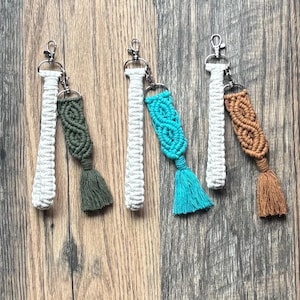 May include: Four macrame keychains in white, green, turquoise, and brown. Each keychain has a woven design and a tassel. The keychains are attached to silver-colored metal clasps. The background is a wooden surface.