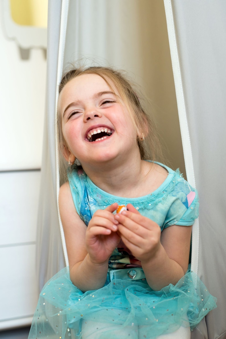 May include: A young girl wearing a light blue dress with a sparkly tulle skirt laughs while sitting in a white fabric swing.
