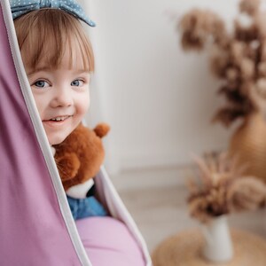 Puede incluir: Una ni&ntilde;a peque&ntilde;a con cabello rubio y ojos azules sonr&iacute;e mientras est&aacute; sentada en una silla colgante morada. Ella est&aacute; sosteniendo un oso de peluche marr&oacute;n.