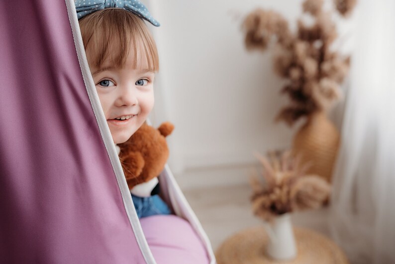 Puede incluir: Una ni&ntilde;a peque&ntilde;a con cabello rubio y ojos azules sonr&iacute;e mientras est&aacute; sentada en una silla colgante morada. Ella est&aacute; sosteniendo un oso de peluche marr&oacute;n.
