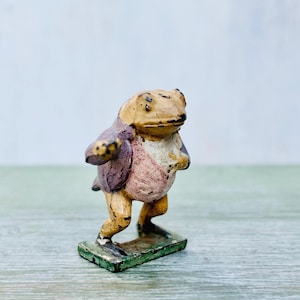 以下が含まれることがあります： 紫とピンクのジャケットを着たカエルの小さな手描きフィギュア。カエルは緑色の土台の上に立っています。
