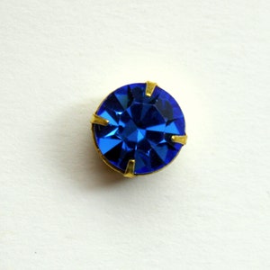 May include: A single, round, blue rhinestone with gold-toned metal settings.