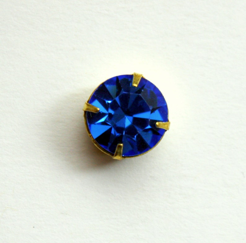 May include: A single, round, blue rhinestone with gold-toned metal settings.