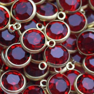 May include: A close-up of a pile of red glass gems with gold metal settings. The gems are round and have a faceted design. The gems are all the same size and are arranged in a random pattern.