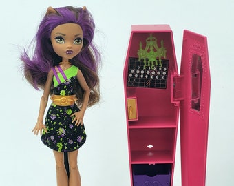 Könnte beinhalten: Ein rosa Sarg-förmiges Puppenhaus mit einer violetten Schublade und einem grünen Kronleuchter im Inneren. Eine Puppe mit violettem Haar und einem schwarzen und grünen Kleid steht neben dem Puppenhaus.
