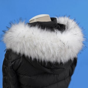 Op de afbeelding: Een zwarte pufferjas met een witte nep bontkraag. De bont is zacht en pluche, en de jas is warm en stijlvol.