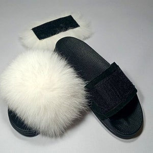 May include: White faux fur slides with a black leather strap and a black suede patch. The slides have a black leather sole.
