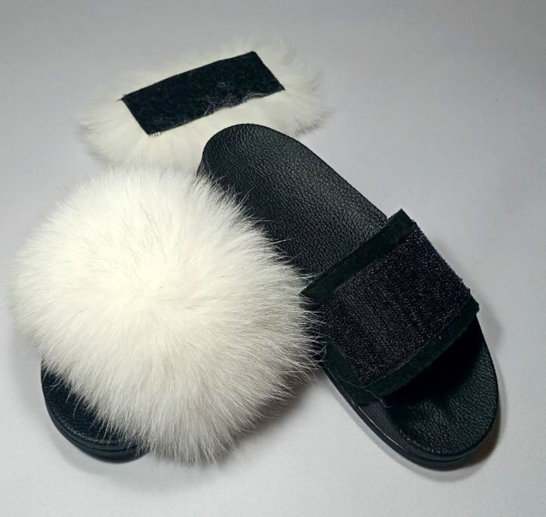 May include: White faux fur slides with a black leather strap and a black suede patch. The slides have a black leather sole.