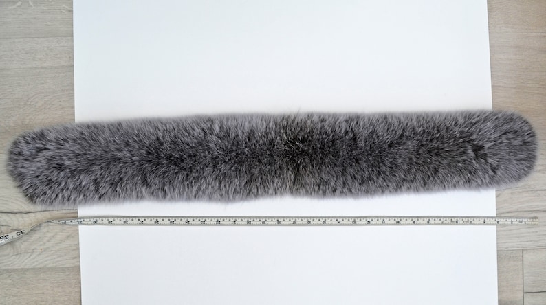 K&ouml;nnte beinhalten: Ein langer, grauer Pelzschal mit weicher, flauschiger Textur. Der Schal wird mit einem Ma&szlig;band gemessen, um seine L&auml;nge zu zeigen.