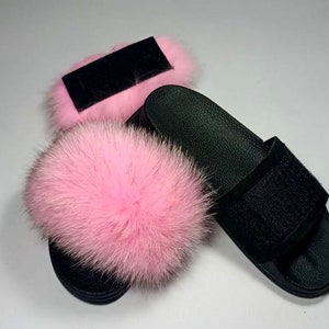 May include: A pair of black leather slides with a pink fur pom-pom on each strap. The slides have a black velcro strap across the top.