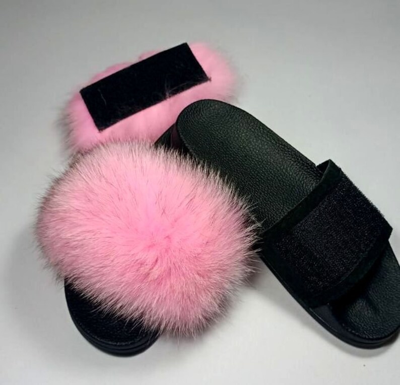 May include: A pair of black leather slides with a pink fur pom-pom on each strap. The slides have a black velcro strap across the top.