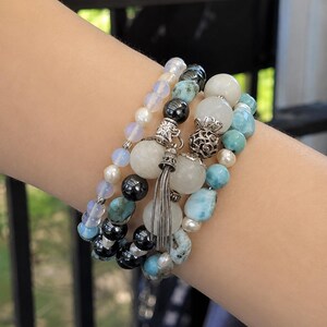 May include: A multi-strand bracelet set featuring a combination of smooth white moonstone beads, iridescent opal-like beads, polished black hematite spheres, and textured light blue turquoise-colored stones, accented with silver-toned metal charms.