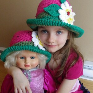 May include: A young girl wearing a pink and green crocheted strawberry hat with a white daisy and ladybug embellishment. She is holding a doll wearing a similar hat.