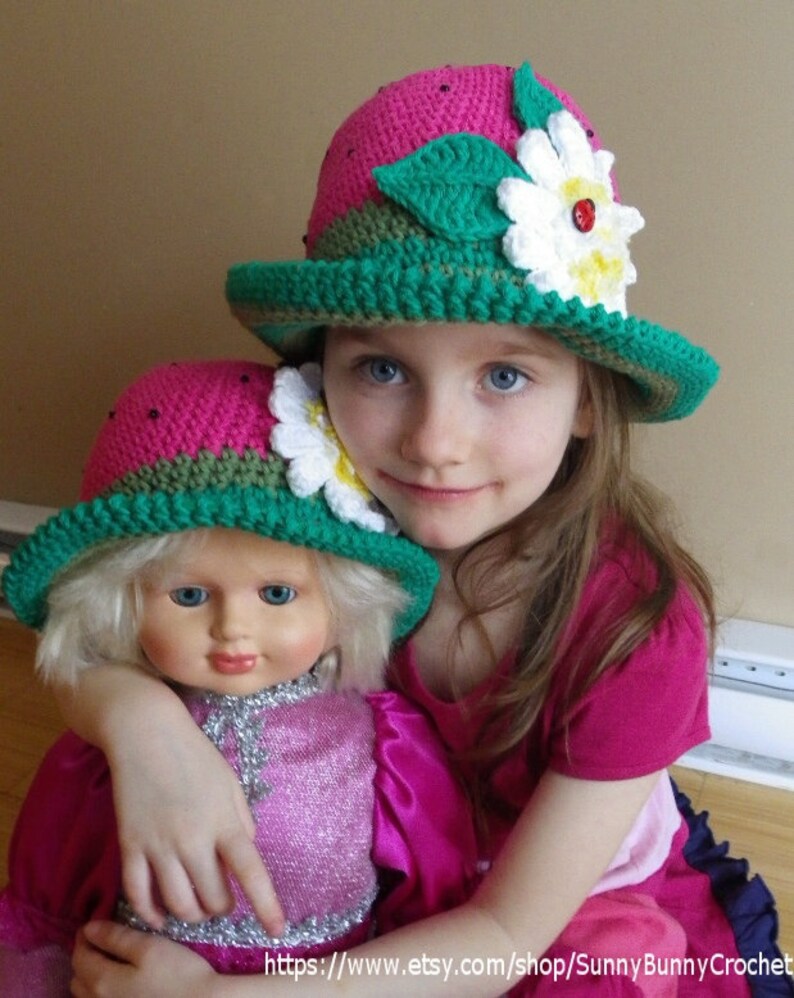 May include: A young girl wearing a pink and green crocheted strawberry hat with a white daisy and ladybug embellishment. She is holding a doll wearing a similar hat.