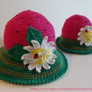 May include: Two crocheted watermelon hats with green and brown stripes, a white flower with a yellow center and a ladybug, and green leaves. The hats are pink and have black beads on the top.