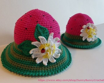 May include: Two crocheted watermelon hats with green and brown stripes, a white flower with a yellow center and a ladybug, and green leaves. The hats are pink and have black beads on the top.
