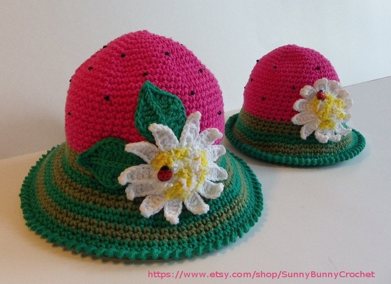 May include: Two crocheted watermelon hats with green and brown stripes, a white flower with a yellow center and a ladybug, and green leaves. The hats are pink and have black beads on the top.