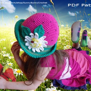 May include: A pink and green crocheted watermelon hat with a white flower and green leaves. The hat is worn by a child lying in a field of daisies.