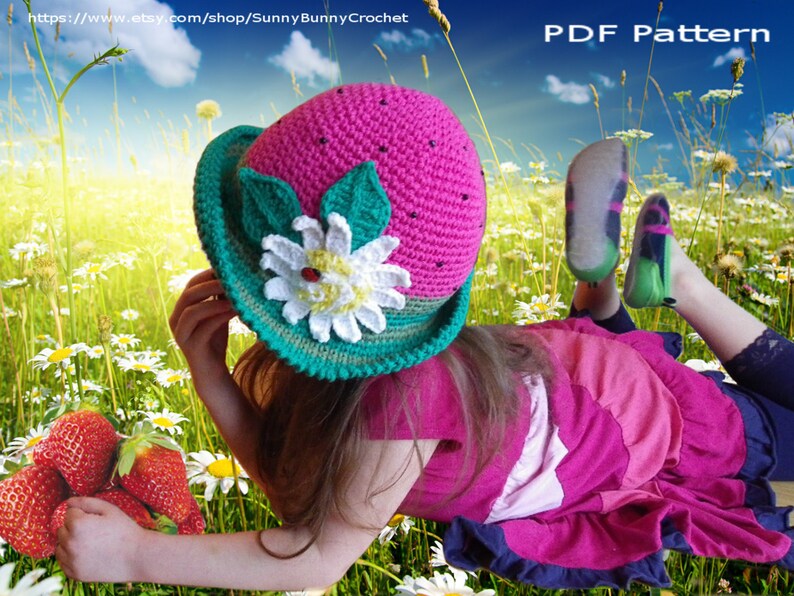 May include: A pink and green crocheted watermelon hat with a white flower and green leaves. The hat is worn by a child lying in a field of daisies.