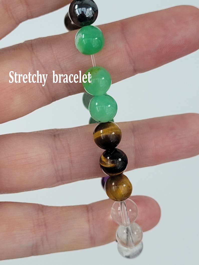 May include: A stretchy bracelet featuring a variety of colorful beads. The beads include green, brown, clear, and tiger's eye stones. The bracelet is held in a hand against a light background.