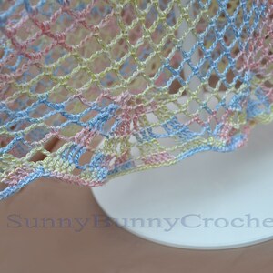 May include: A close-up of a crocheted fabric with a light blue, pink, and yellow color scheme. The fabric has a delicate, open-weave pattern.