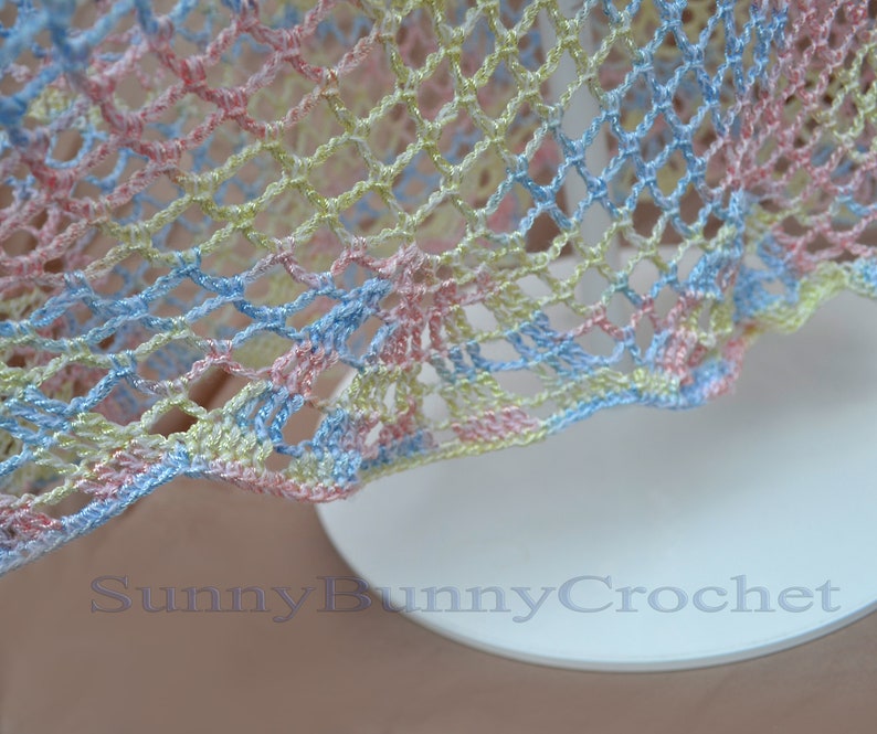 May include: A close-up of a crocheted fabric with a light blue, pink, and yellow color scheme. The fabric has a delicate, open-weave pattern.