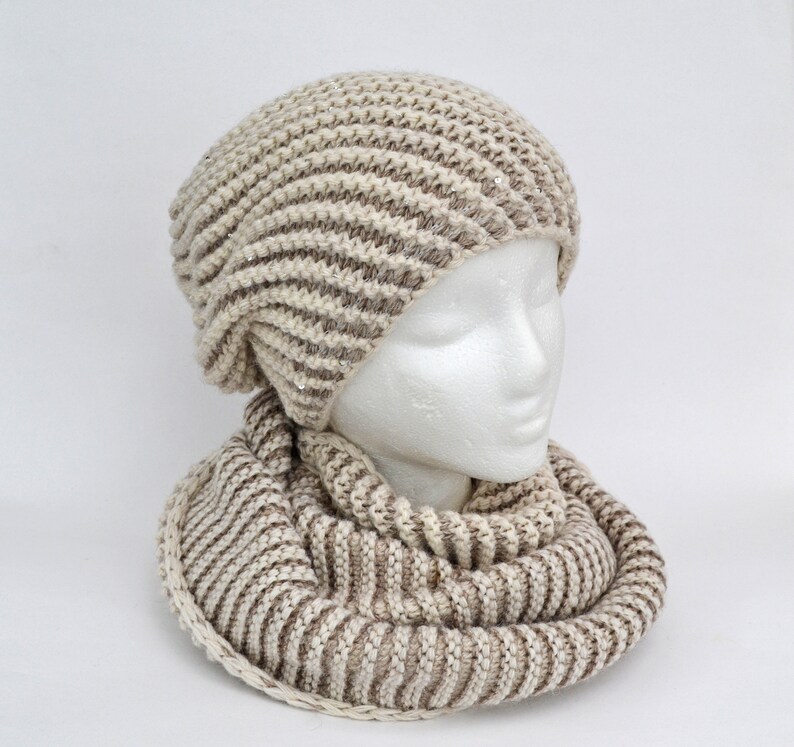 May include: A beige and brown striped knit hat and matching scarf. The hat has a slouchy fit and the scarf is a cowl style.