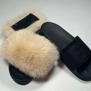 May include: A pair of black faux fur slides with a black suede strap. The slides have a fluffy, light brown fur pom-pom on the top.