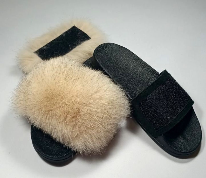 May include: A pair of black faux fur slides with a black suede strap. The slides have a fluffy, light brown fur pom-pom on the top.