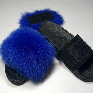May include: A pair of black faux fur slides with blue fox fur pom poms. The slides have a black strap with a velcro closure.