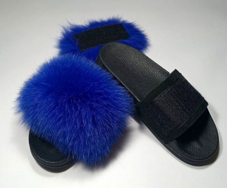 May include: A pair of black faux fur slides with blue fox fur pom poms. The slides have a black strap with a velcro closure.